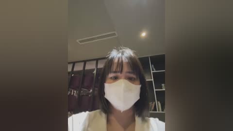 Media: A video of a woman with straight, shoulder-length black hair, wearing a white face mask and a white blouse, standing in a dimly lit room with a window and a closet in the background.