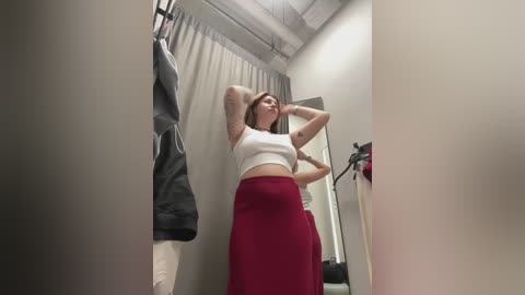 Media: Video of a young woman in a dressing room, adjusting her white crop top and red skirt, standing in front of beige curtains and a mirror.