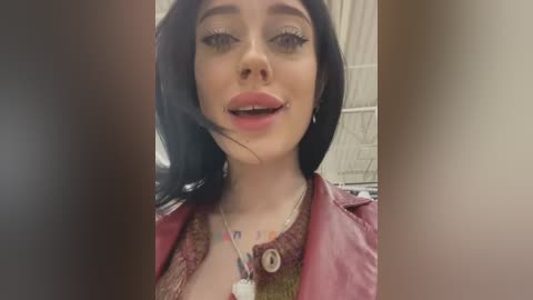 Media: Video of a young woman with fair skin, long black hair, and blue eyes, wearing a maroon leather jacket, a beaded necklace, and a silver nose ring. She has a slight smile and a tattoo visible on her neck.