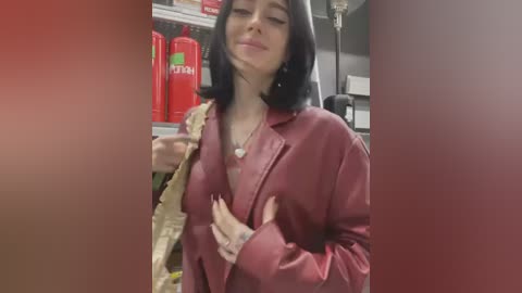 Media: A video of a woman with shoulder-length black hair and fair skin, wearing a maroon jacket, holding a plastic bag with a smile. Background includes red and green soda bottles and shelves.