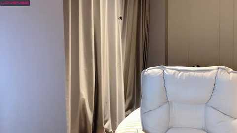 Media: Video of a minimalist room with beige curtains covering a window, a white cushioned chair, and a small, framed painting in the top left corner.