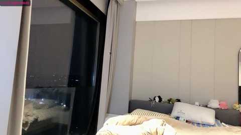 Media: Video of a modern bedroom with beige walls, a large window with a city skyline view, a bed with white and beige sheets, and various decorative items on the bedside table.