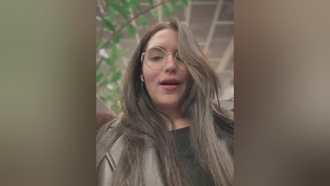 Media: Video of a young woman with long, straight brown hair, wearing glasses and a black shirt, standing indoors near a window with greenery outside.