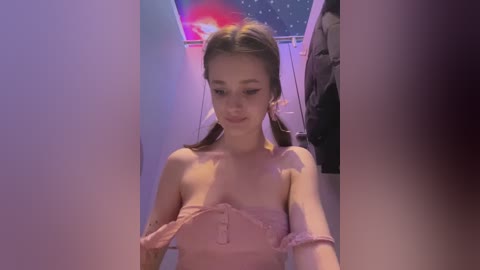 Media: Video of a young Caucasian woman with straight brown hair in pigtails, wearing a strapless pink dress, standing in a narrow, dimly lit room with a starry sky-themed wall.