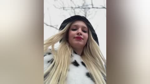 A video of a young blonde woman with long, straight hair in a black hat, wearing a white and black cow-patterned jacket, standing in a snowy outdoor setting with bare tree branches in the background.