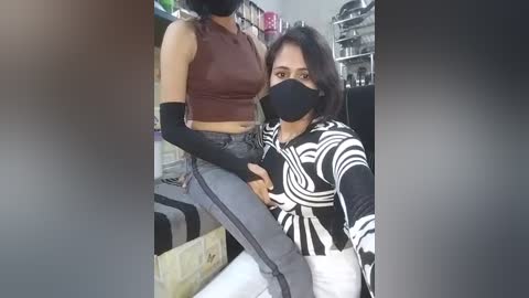 Video of two young women, one with long dark hair and a mask, wearing a black top with white patterns, sitting on a counter, while the other, with short hair, wearing a brown crop top and grey pants, stands behind.