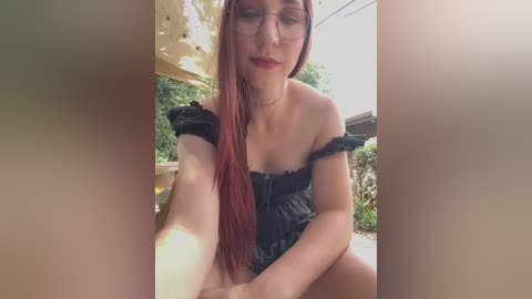 Media: Video of a fair-skinned woman with long red hair and glasses, wearing a black off-shoulder dress, sitting outdoors under a yellow canopy, with a blurred garden background.