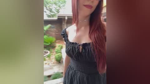 Media: A video of a fair-skinned woman with long, straight red hair, wearing a black off-shoulder dress, standing in a garden with green plants and a wooden fence background.