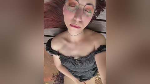 Media: Video of a fair-skinned, curly-haired woman with glasses and green makeup on her eyes, lying on a wooden surface in a black off-shoulder dress, with her legs slightly apart.