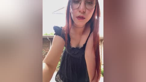 Media: Video of a young woman with long red hair, wearing glasses, a black lace top, and standing outdoors. The background shows a garden and a brick building.