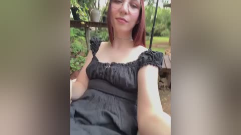 Media: A video of a young, fair-skinned woman with straight red hair, wearing a black ruffled top and sitting outdoors in a garden. Background features greenery, potted plants, and a wooden structure.