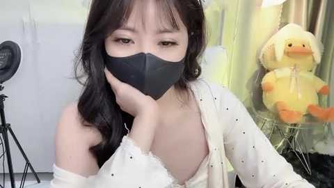 Media: Video of an East Asian woman with long black hair and a black face mask, wearing a white, off-shoulder top, sitting in front of a yellow duck toy and a camera on a tripod.