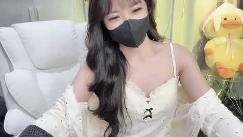 Media: Video of an East Asian woman with long, wavy black hair, wearing a white lace dress, black face mask, and black gloves, sitting in a white chair in a room with plush toys and a green wall.