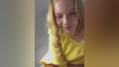 Media: Video of a young, fair-skinned, blonde-haired woman with a soft smile, wearing a yellow off-shoulder top, with a blurred background.