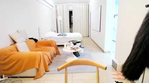 Media: Video of a minimalist, modern hotel room with beige walls, light wooden furniture, and a queen-sized bed covered in white linens. An orange throw blanket and pillows adorn a beige sofa. A wooden chair is visible in the foreground.