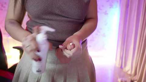 Media: Video of a woman in a gray ribbed top and white skirt, holding a white, fluffy object. Background features a pastel-colored, softly lit room with floral wallpaper and curtains.