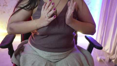 Media: Video of a plus-sized woman with long dark hair, wearing a brown ribbed tank top and gray skirt, seated in a black office chair, hands pressed to her chest, with purple nails.