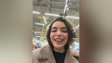 Video of a young woman with light brown skin, straight dark hair, and braces, smiling brightly, wearing a tan coat and black top inside a brightly lit, modern grocery store with high ceilings and fluorescent lighting.