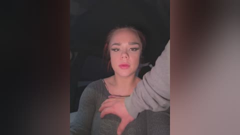 Media: Video of a young woman with light skin, brown hair in a ponytail, wearing pink lipstick, sitting in a dimly lit car, with a man's hand on her shoulder.