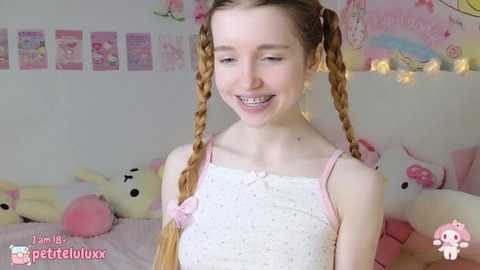 Media: A video of a smiling, pale-skinned, young woman with long braided hair in a white polka-dot dress, surrounded by plush toys in a pink, whimsical room.