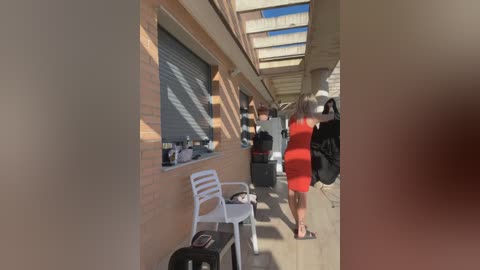 Media: Video of a woman in a red dress walking away from a brick building with a roll-up window, next to a white plastic chair and black suitcase on the sunlit sidewalk.