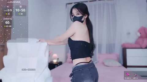 Media: Video of a young Asian woman with long black hair, wearing a black mask and crop top, in a pink bedroom.