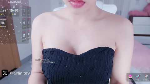 Video of an Asian woman with fair skin, wearing a black strapless dress, standing in a modern room with a pink bed and white walls.