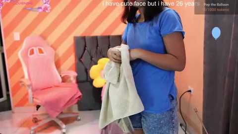 A video of a woman in a blue shirt and jeans holding a yellow towel in a vibrant, orange-striped room with a pink gaming chair and a black couch.