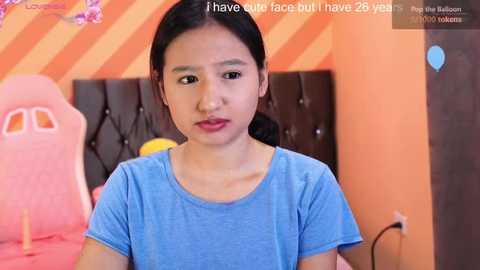 Video of an Asian girl with straight black hair, fair skin, wearing a blue t-shirt, sitting in an orange-striped room with a pink gaming chair.