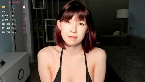 Media: Video of a young Asian woman with straight, shoulder-length dark brown hair, fair skin, and a slim physique, wearing a black halter top. She stands in a modern, dimly lit living room with a grey sofa and a white lamp.