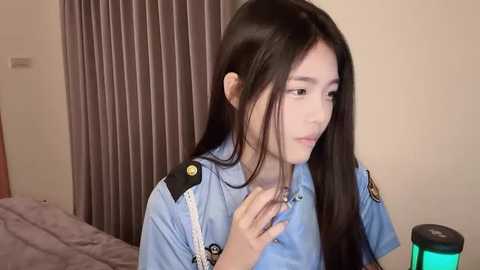 Media: Video of an East Asian woman with long black hair, wearing a light blue police uniform, looking contemplative in a beige room with vertical blinds and a bed in the background.