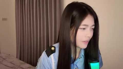 Media: Video of a young East Asian woman with long black hair, wearing a light blue shirt, looking at a phone, in a beige room with vertical blinds.