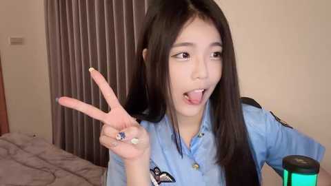 Media: Video of an East Asian woman with straight black hair and fair skin, wearing a light blue shirt, making a peace sign with her right hand. She's in a bedroom with a bed and beige walls.