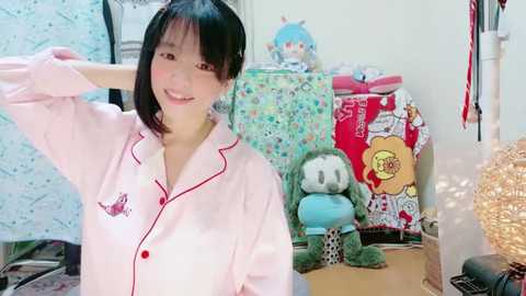 Media: Video of an East Asian woman with short black hair, wearing a pink pajama shirt with a cartoon character, smiling in a cluttered bedroom with stuffed toys and a floral bedspread.