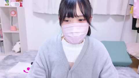 Media: A video of a young Asian woman with dark hair, wearing a light gray robe and pink face mask, in a softly lit, minimalist bedroom with white walls and pastel decor.