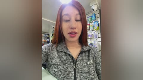 Media: Video of a young, light-skinned Asian woman with straight, shoulder-length auburn hair, wearing a gray zip-up hoodie, smiling softly. Background features a pharmacy with shelves of various medications.