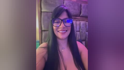 Media: Video of a smiling, fair-skinned woman with long black hair, wearing black-rimmed glasses and a black top, standing in a dimly lit room with a stone wall and purple lighting.