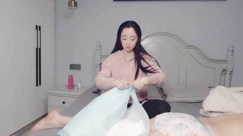 Media: Video of an Asian woman with long black hair, wearing a pink sweater, ironing a light blue shirt in a white bedroom with a white bed and dresser.