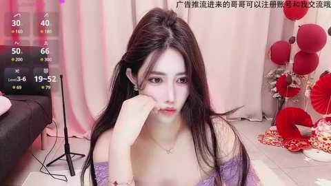 Media: Video of an Asian woman with long black hair, wearing a purple off-shoulder dress, sitting indoors with a sad expression. Background features red and white decorations, pink curtains, and a TV screen displaying a temperature of 40\u00b0C.