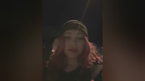 Media: A dimly lit video of a young woman with long, wavy red hair, wearing a dark beanie and black shirt, leaning forward, eyes closed, and lips slightly parted. The background is dark and blurry.