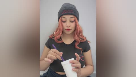 Media: Video of a young woman with long, wavy, pastel pink hair, wearing a gray beanie, black crop top, and blue jeans, writing on a white paper with a purple pen.