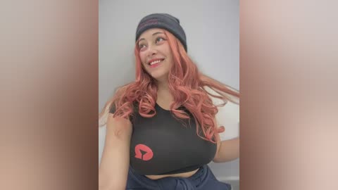 Media: Video of a smiling woman with long, wavy, pink hair, wearing a black crop top with a red heart and navy blue pants, against a plain white background.