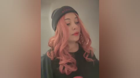 Media: Video of a young woman with light skin, wavy pink hair, and a dark beanie, wearing a black sweater. Soft, blurred background.