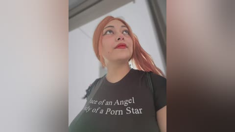 Media: Video of a young woman with fair skin and long, pink hair, wearing a black t-shirt with white text, looking upward in an indoor setting.