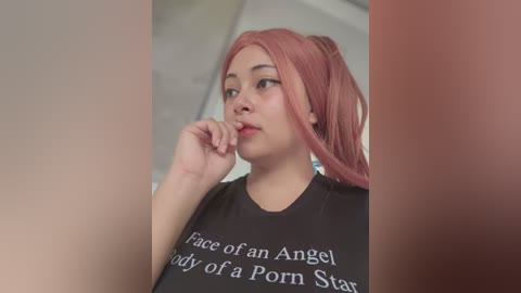 Media: Video of an Asian woman with long, pastel pink hair in a ponytail, wearing a black t-shirt with the phrase \"Face of an Angel, body of a Porn Star.\