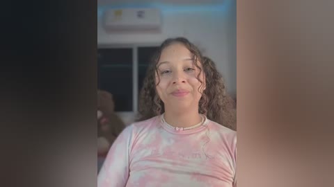 Media: Video of a light-skinned woman with curly hair, wearing a tie-dye shirt, standing indoors with a blurred background.