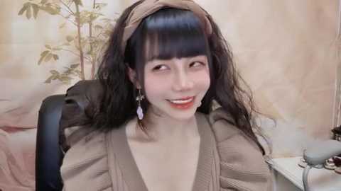 Media: Video of an Asian woman with long, dark hair, wearing a beige blouse, sitting in an office chair with a beige headband. Background features floral wallpaper and a phone.