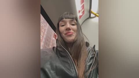 Media: Video of a young woman with long, straight, dark hair, wearing a black leather jacket, standing in a subway car. She has a neutral expression and is partially obscured by the frame.