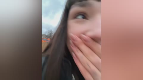 Media: Video of a close-up of a woman's face with brown hair, partially obscured by her hand, taken outdoors in daylight. Background shows a blurred urban setting with buildings.