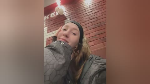 Media: Video of a young woman with light skin, long blonde hair, and a gray beanie, wearing a gray jacket with a hexagonal pattern. She leans against a red brick wall with white trim and a vintage-style light fixture.